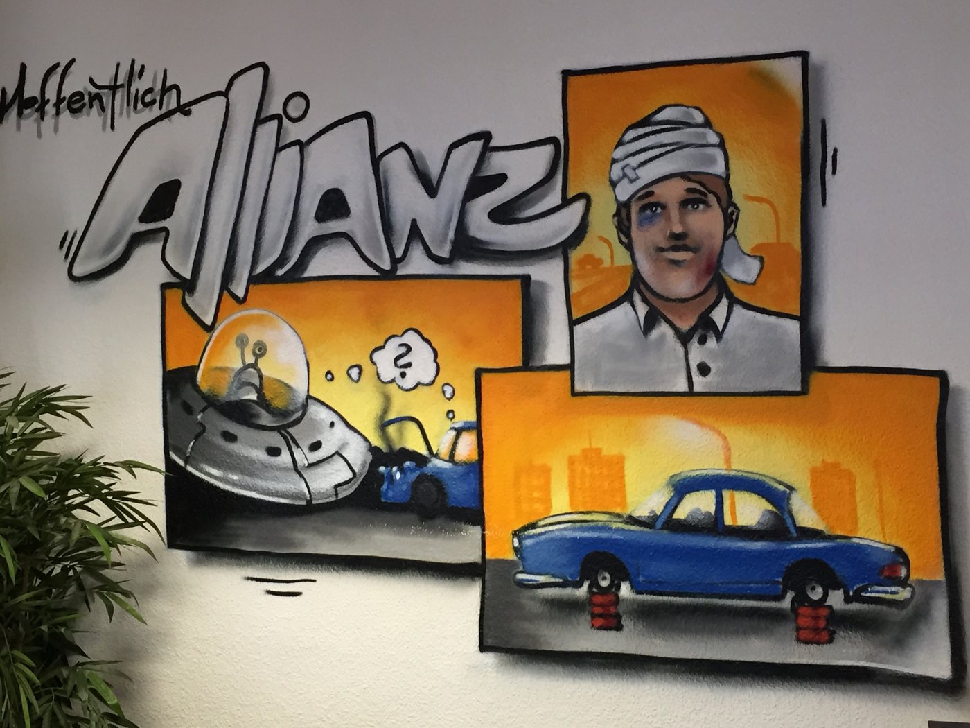 Graffiti an der Wand der Agentur: Schriftzug Allianz, daneben ein Ufo, das auf ein Auto gefallen ist, ein Mann mit Kopfverband und ein Auto ohne Räder